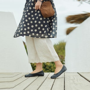 Puede incluir: Bailarinas de cuero negro. Los zapatos los lleva una persona que viste un vestido azul marino con lunares y pantalones color crema. La persona está de pie en una terraza de madera.