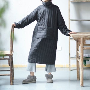 May include: A black quilted tunic dress with a large patch pocket on the front. The dress has a mock neck and is worn with gray pants and gray fuzzy boots.