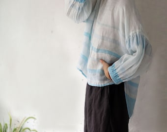 Puede incluir: Una camisa de lino de rayas blancas y azul claro, de corte holgado y manga larga. La camisa tiene un detalle fruncido en los puños y un dobladillo ligeramente recortado.