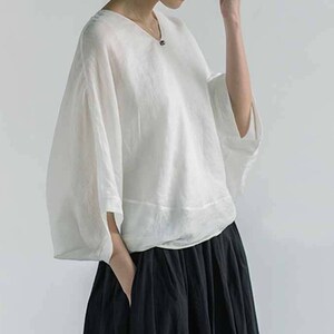 May include: A white linen blouse with a v-neck and 3/4 sleeves. The blouse is loose fitting and has a relaxed silhouette. The model is wearing a black skirt.