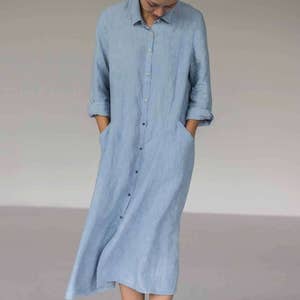 Pode incluir: Vestido camisa de linho azul claro com mangas compridas e fecho de botões. O vestido tem dois bolsos frontais e uma gola de camisa. As mangas estão enroladas. O vestido é até ao joelho.
