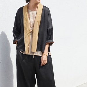May include: A black and tan linen jacket with a long, open front and wide sleeves. The jacket is worn over a white tank top and black cropped pants. The person is wearing black sandals.