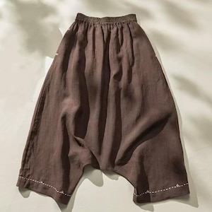May include: Brown linen pants with a gathered waistband and wide legs. The pants have a relaxed fit and feature white stitching details along the hem. The fabric has a slightly wrinkled texture.