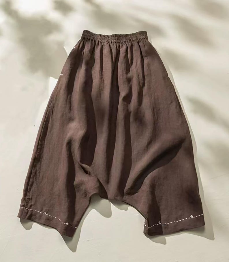 May include: Brown linen pants with a gathered waistband and wide legs. The pants have a relaxed fit and feature white stitching details along the hem. The fabric has a slightly wrinkled texture.