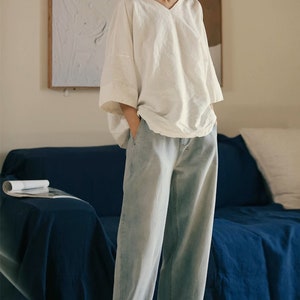 May include: A white linen top with a v-neck and 3/4 sleeves. The top is loose fitting and has a relaxed silhouette. The model is wearing the top with light blue jeans.