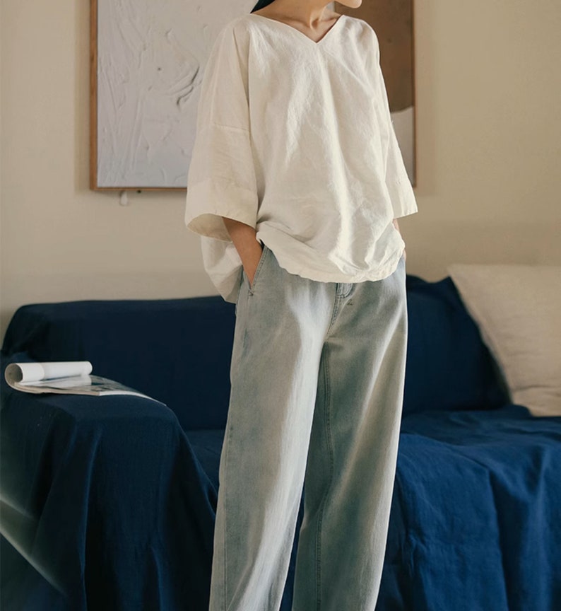 May include: A white linen top with a v-neck and 3/4 sleeves. The top is loose fitting and has a relaxed silhouette. The model is wearing the top with light blue jeans.