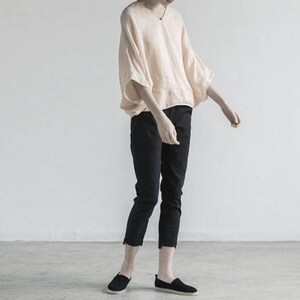 May include: A person wearing a loose-fitting, light peach colored linen top with a wide, flowing neckline and 3/4 sleeves. They are wearing black cropped pants and black slip-on shoes.