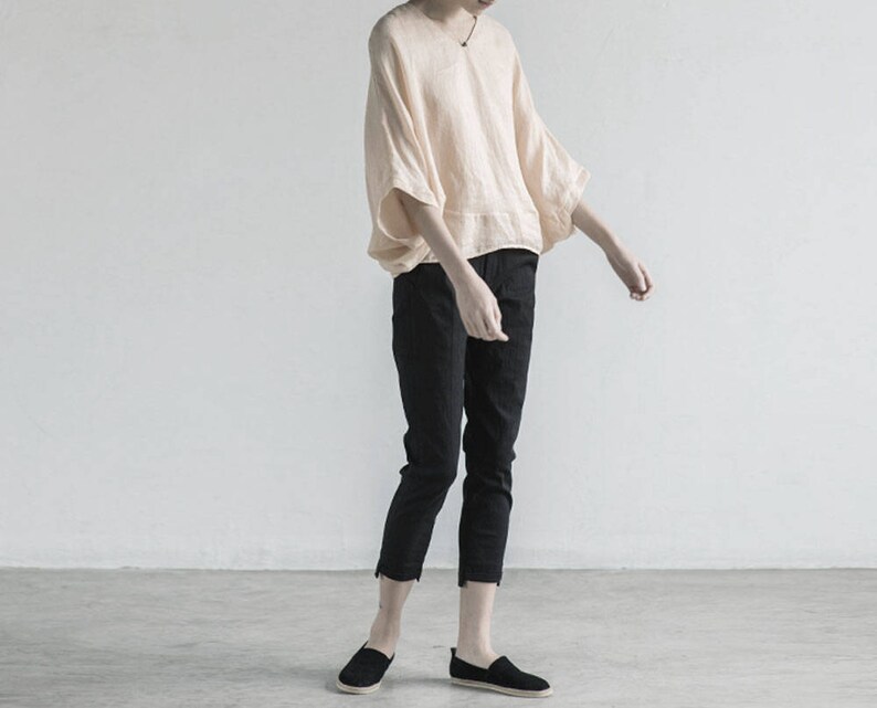 May include: A person wearing a loose-fitting, light peach colored linen top with a wide, flowing neckline and 3/4 sleeves. They are wearing black cropped pants and black slip-on shoes.