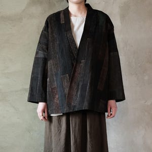 May include: A dark, patchwork-style jacket with a V-neck design. The jacket features a mix of black, brown, and gray rectangular panels. The sleeves are wide and the jacket is worn over a white top and brown skirt.