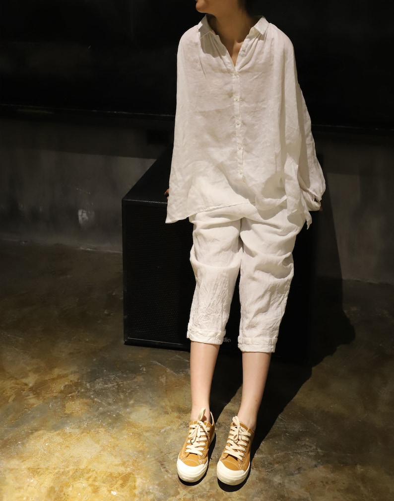 Pu&ograve; includere: Una camicia e pantaloni di lino bianco con una vestibilit&agrave; rilassata. La camicia ha un colletto abbottonato e maniche lunghe. I pantaloni sono corti e hanno una gamba larga. La persona indossa sneakers di tela marroni.