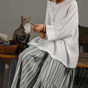 May include: A woman wearing a white linen top with a silver necklace and a striped skirt. She is sitting on a wooden bench and holding a green leaf in her hand. A tabby cat is sitting next to her.