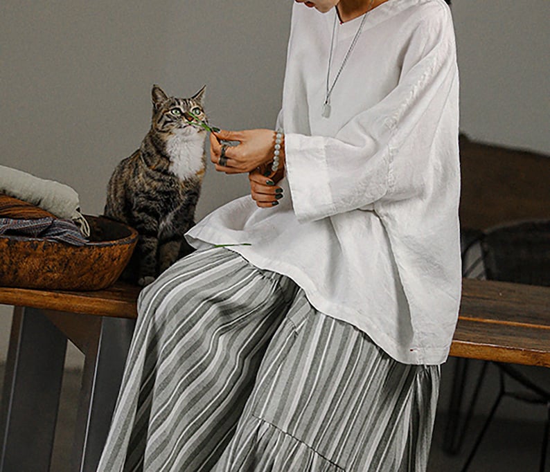 May include: A woman wearing a white linen top with a silver necklace and a striped skirt. She is sitting on a wooden bench and holding a green leaf in her hand. A tabby cat is sitting next to her.