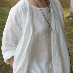 May include: A white linen top with a long, flowing sleeve and a simple, round neckline. The top is layered over a sheer white shirt, creating a relaxed and elegant look.