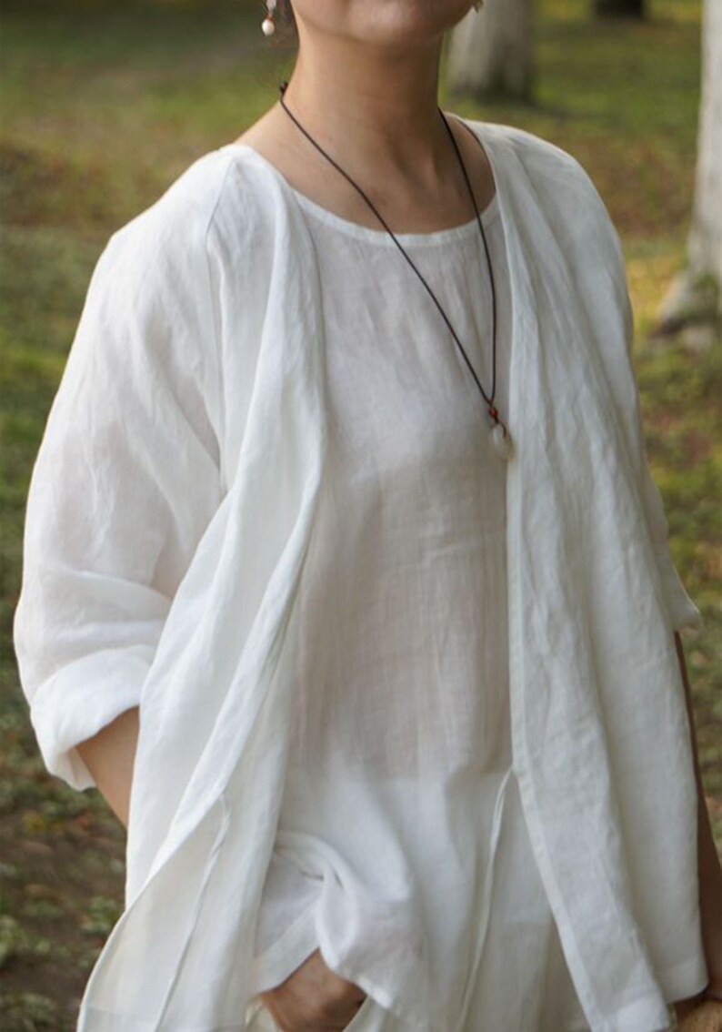 May include: A white linen top with a long, flowing sleeve and a simple, round neckline. The top is layered over a sheer white shirt, creating a relaxed and elegant look.