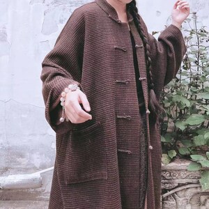 May include: A brown long-sleeved jacket with a mandarin collar and button closures. The jacket has a textured woven fabric and a single pocket on the left side.