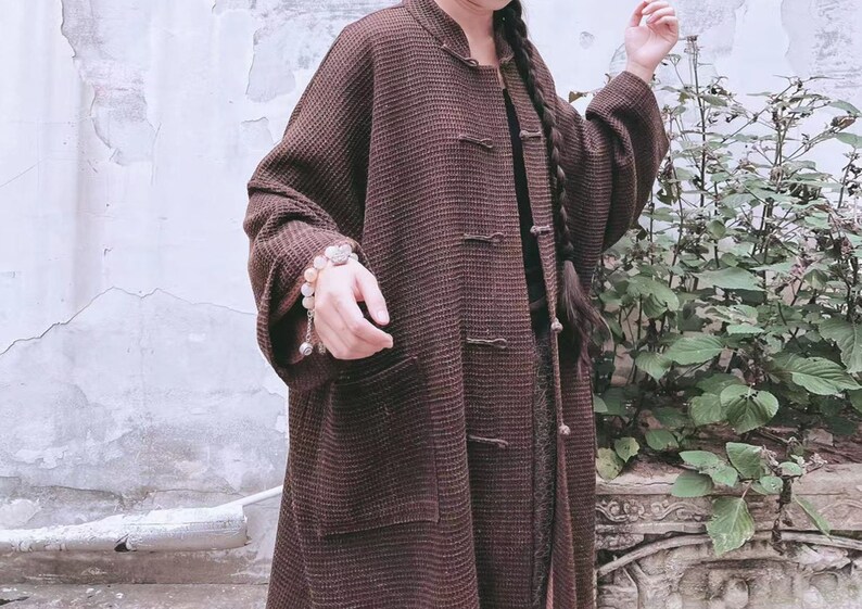 May include: A brown long-sleeved jacket with a mandarin collar and button closures. The jacket has a textured woven fabric and a single pocket on the left side.