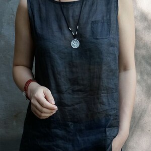 May include: A sleeveless black linen top with a small pocket on the left side. The model is wearing a silver necklace with a round pendant.