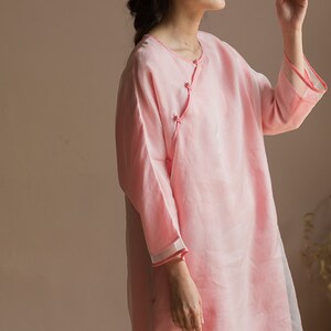 Op de afbeelding: Een lichtroze, langarmige, losse tuniek met een knoopsluiting aan de linkerkant. De tuniek heeft een eenvoudig, elegant ontwerp en is gemaakt van een lichtgewicht stof.