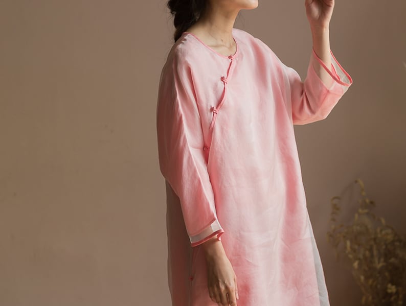 Op de afbeelding: Een lichtroze, langarmige, losse tuniek met een knoopsluiting aan de linkerkant. De tuniek heeft een eenvoudig, elegant ontwerp en is gemaakt van een lichtgewicht stof.