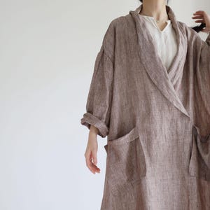 Pode incluir: Um roupão longo e fluido de linho castanho com grandes bolsos frontais e gola de xale. O roupão é usado sobre um top branco. As mangas são enroladas nos cotovelos. O roupão é feito de um tecido texturizado.