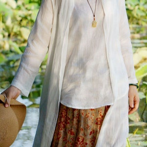 May include: A white linen long-sleeved shirt with a matching white linen cardigan. The cardigan is open in the front and has a long, flowing design. The shirt has a simple, elegant style. The person is wearing a brown and white patterned skirt.