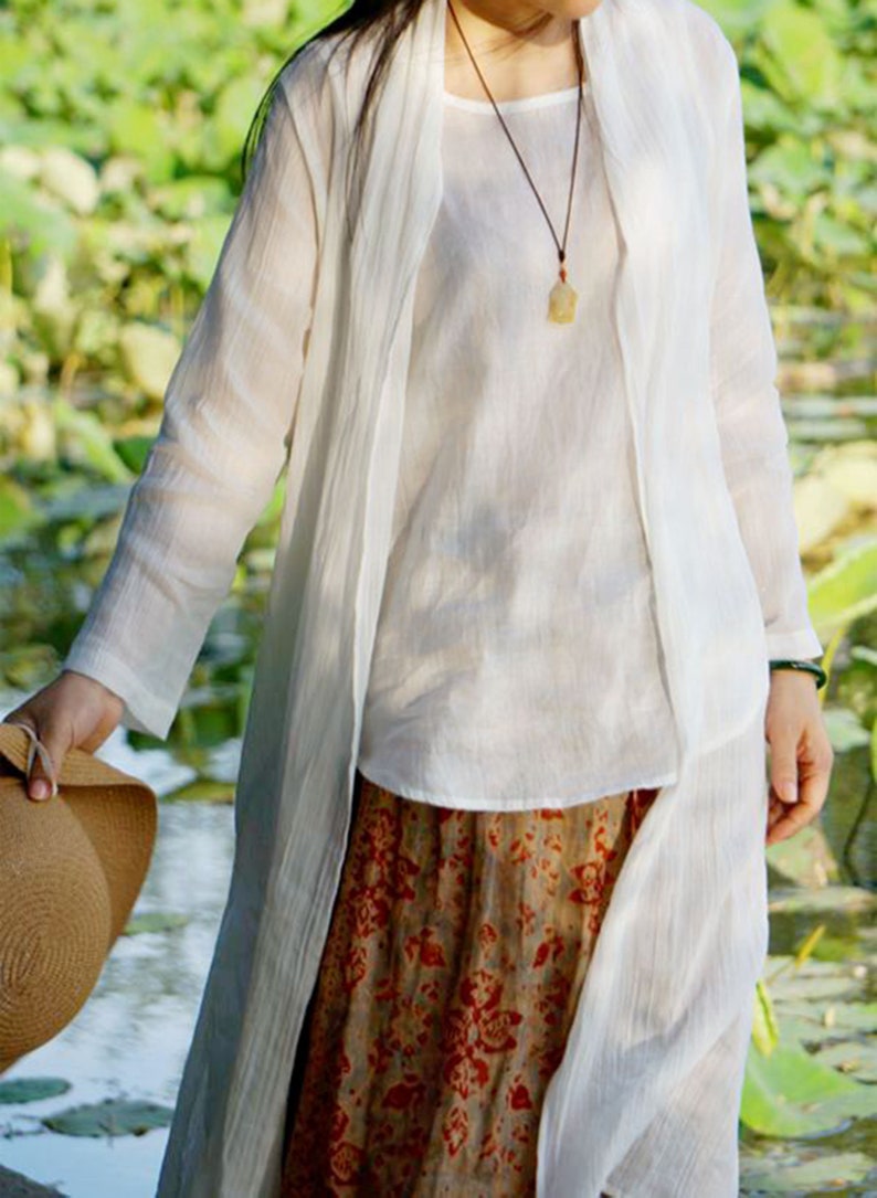 May include: A white linen long-sleeved shirt with a matching white linen cardigan. The cardigan is open in the front and has a long, flowing design. The shirt has a simple, elegant style. The person is wearing a brown and white patterned skirt.