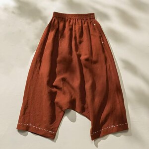May include: Rust-colored linen pants with a relaxed fit. The pants have a gathered waistband and a drop-crotch design. White stitching accents the hemline. The fabric appears lightweight and breathable, suitable for warm weather.
