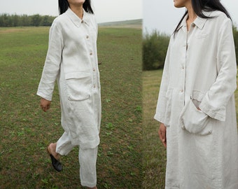 May include: A long, white linen coat with a button-up front and a large pocket on the left side. The coat is worn over a pair of white linen pants.