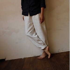 May include: A pair of white linen trousers with a wide leg and a relaxed fit. The trousers are cuffed at the bottom and are being worn by a person standing against a wall.