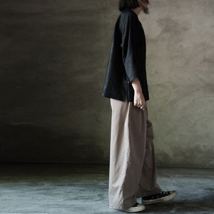 Puede incluir: Una persona lleva una camisa negra de manga larga y pantalones anchos de color marr&oacute;n claro. Est&aacute; de pie frente a una pared gris texturizada.