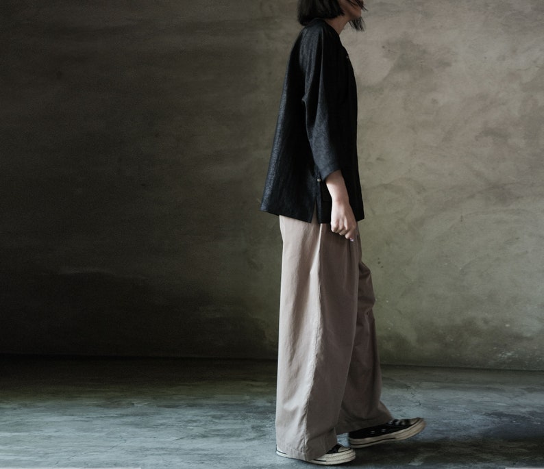 Puede incluir: Una persona lleva una camisa negra de manga larga y pantalones anchos de color marr&oacute;n claro. Est&aacute; de pie frente a una pared gris texturizada.