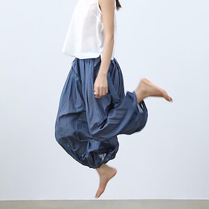 以下が含まれることがあります： 白いノースリーブのトップスと青いハーレムパンツを着用した人。パンツは足首でギャザーが寄せられ、ボリュームのあるシルエットを作り出しています。片足を伸ばし、ジャンプの最中です。