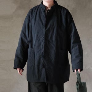 May include: A black, long-sleeved jacket with a mandarin collar and two front pockets. The jacket has a button closure and a relaxed fit. The person is holding a green tote bag. The jacket appears to be made of a lightweight fabric.