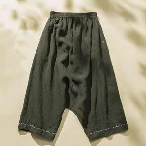 May include: Dark olive green linen pants with a relaxed fit. The pants have an elastic waistband and white stitching details along the hem and side seams. The fabric has a slightly wrinkled texture, suggesting a lightweight and breathable material.