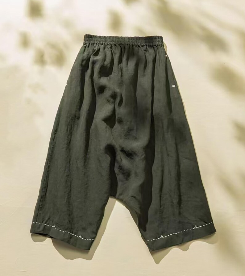 May include: Dark olive green linen pants with a relaxed fit. The pants have an elastic waistband and white stitching details along the hem and side seams. The fabric has a slightly wrinkled texture, suggesting a lightweight and breathable material.