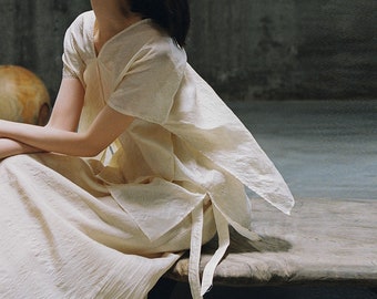 May include: A woman wearing a white linen top with a flowing, asymmetrical design. The top has a V-neck and short sleeves. The woman is sitting on a wooden surface.