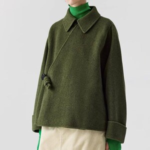 May include: Olive green wrap jacket with a collar and a knot closure. The long-sleeved jacket has a folded cuff and is paired with a green turtleneck and a cream-colored skirt. The jacket is made of a textured fabric.