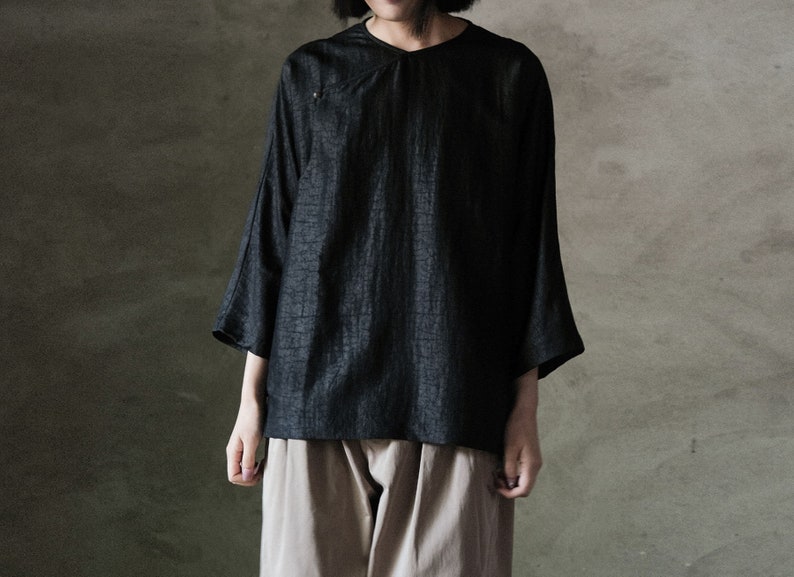 Puede incluir: Una camisa negra holgada de manga 3/4 con cuello redondo. La camisa est&aacute; hecha de una tela texturizada y tiene un ligero efecto fruncido en el cuello.