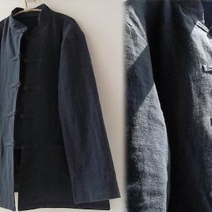 May include: A dark blue jacket with a mandarin collar and frog closures. The jacket has long sleeves and two front pockets. The fabric appears to be a textured linen or cotton blend. The jacket is hanging on a wooden hanger against a white wall.