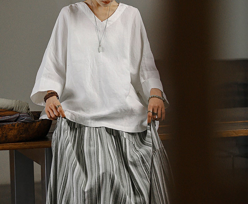 May include: A white linen blouse with a v-neck and 3/4 sleeves paired with a striped skirt. The skirt has a tiered design and is made of a lightweight fabric.