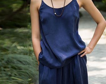 May include: A navy blue camisole top with thin straps and matching wide-leg pants. A long beaded necklace with a white pearl pendant is worn. The outfit is made of a silky material. The model is standing outside in front of a blurred green background.