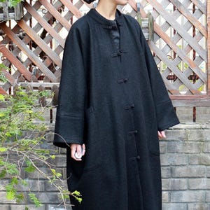 Könnte beinhalten: Ein langer, schwarzer Wollmantel mit Stehkragen und Knebelverschlüssen. Der Mantel hat lange Ärmel und große Taschen. Der Mantel ist knielang und scheint ein Winterkleidungsstück zu sein.