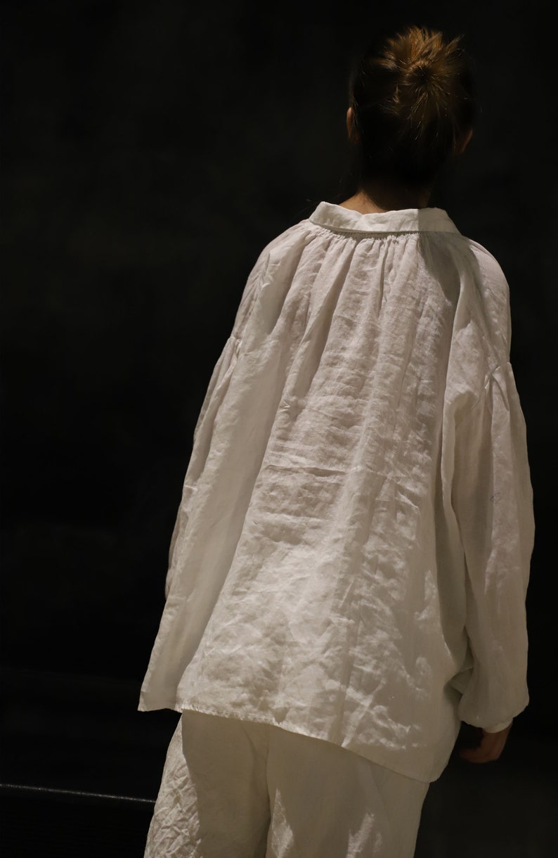 Pu&ograve; includere: Una camicia di lino bianca con una vestibilit&agrave; ampia e oversize e un collo arricciato. La camicia &egrave; indossata sopra pantaloni bianchi.