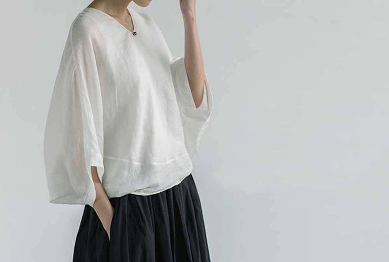 May include: A white linen blouse with a v-neck and 3/4 sleeves. The blouse is loose fitting and has a relaxed silhouette. The model is wearing a black skirt.