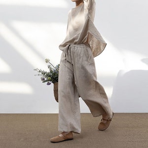 以下が含まれることがあります： ゆったりとした薄いベージュのリネン素材のトップスとそれに合わせたワイドレッグパンツを着用した女性。彼女は花束を持ってカメラに向かって歩いています。