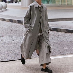 May include: A person wearing a light gray long coat, matching pants, and black shoes. The coat is buttoned and has pockets. The outfit is layered over a white shirt and a sweater. The person is walking on a paved street.