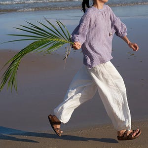 May include: A person wearing a lavender linen top and white linen pants walks on a beach, holding a large green palm frond. The person is wearing brown sandals. The ocean is in the background.