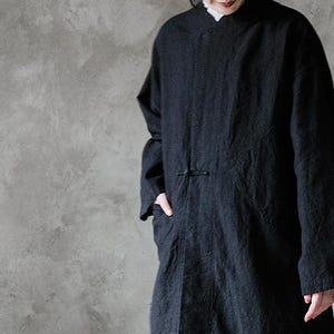 May include: A long, black coat with a mandarin collar and long sleeves. The coat has a simple button closure and pockets. The fabric appears to be a textured linen or cotton blend. The garment is a casual, yet elegant style.