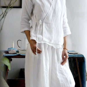 May include: A white linen wrap top with a tie at the waist and matching white linen pants. The top has a relaxed fit and a V-neckline.