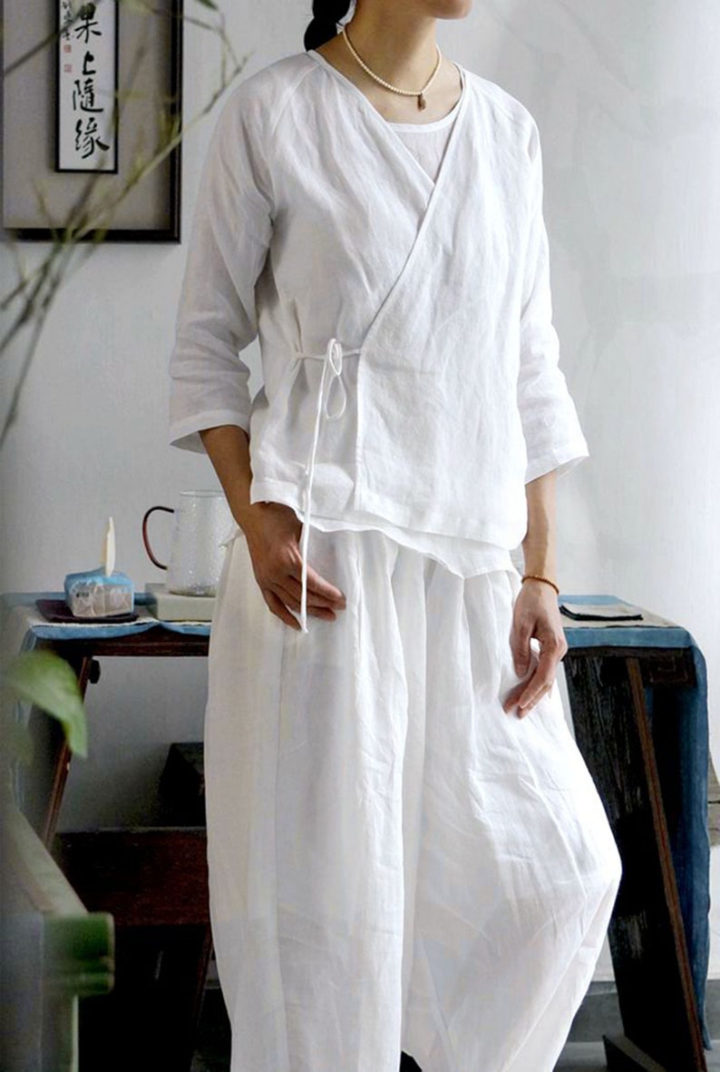 May include: A white linen wrap top with a tie at the waist and matching white linen pants. The top has a relaxed fit and a V-neckline.